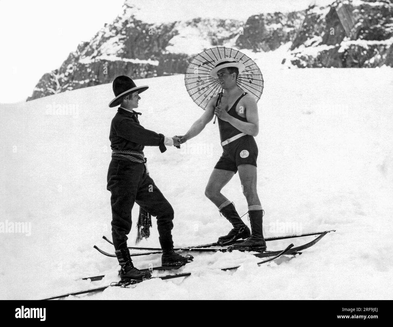 United States: c. 1923. A woman skier on the mountain slope wearing a sash and western hat shakes hands with a man on skis in a one piece bathing suit and carrying a parasol. Stock Photo