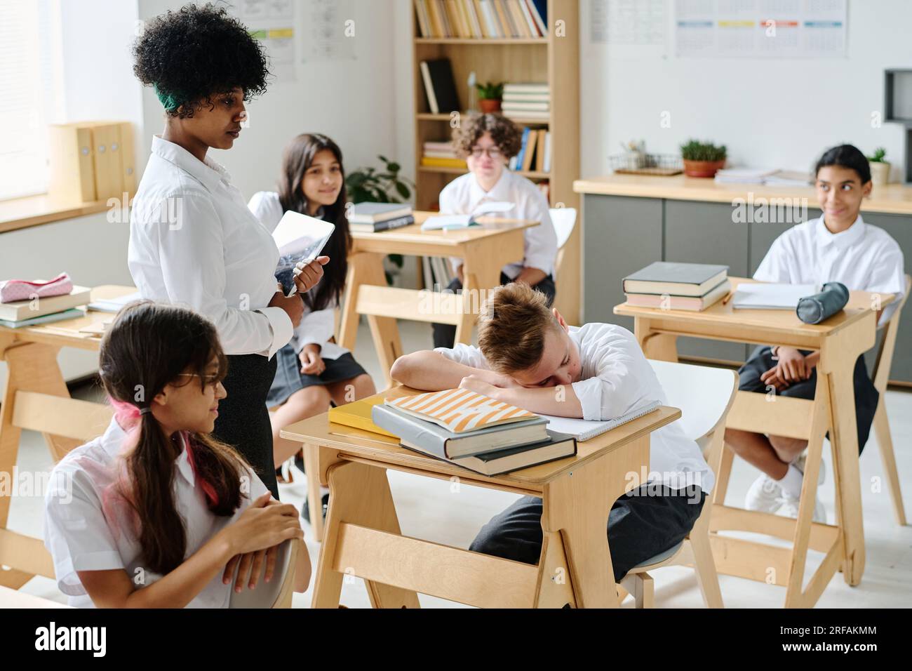 Schoolboy sleeping at his desk during lesson at school while teacher waking him up Stock Photo