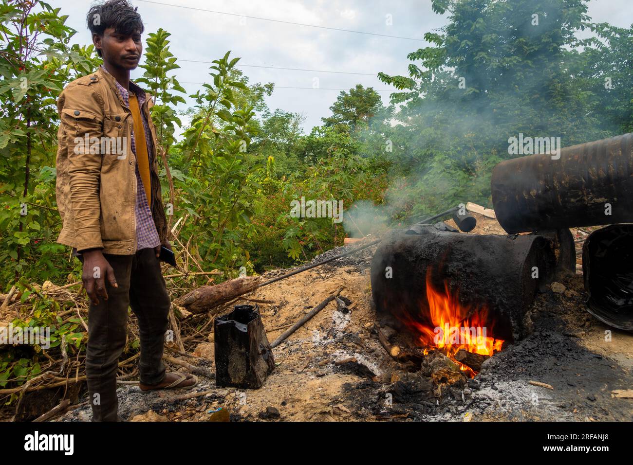 Oct.14th 2022 Uttarakhand, India. Worker igniting charcoal barrels for road construction. Intense flames and billowing black smoke from tar burning. C Stock Photo
