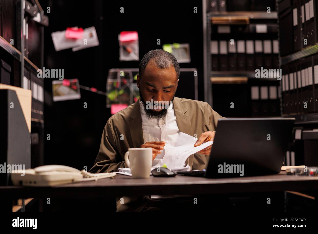 African american police detective searching evidence in crime case file. Private investigator reading witness statement in reports while working overtime and sitting at desk at night time Stock Photo