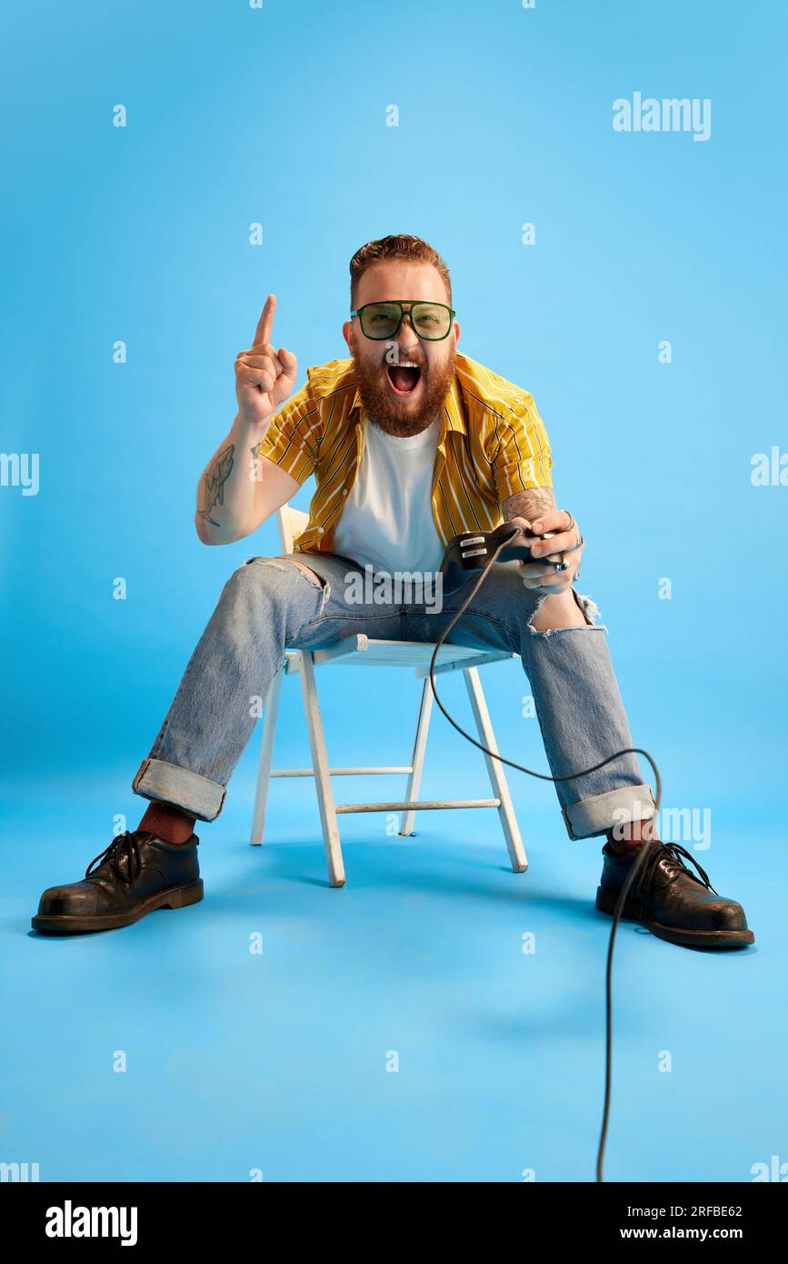Bearded man in casual clothes and sunglasses emotionally playing console against blue studio background. Evening fun Stock Photo