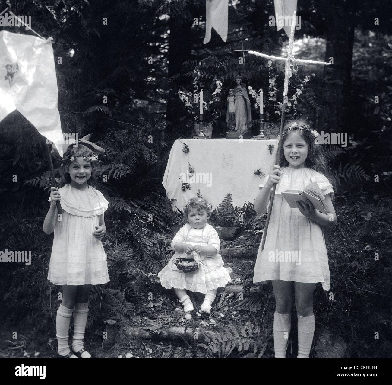 Trois petites filles, en robes de dentelle, portant croix, panier et fanion, devant un autel dedie a la Vierge, en pleine nature. Bretagne (France), photographie 1921-1922. Stock Photo