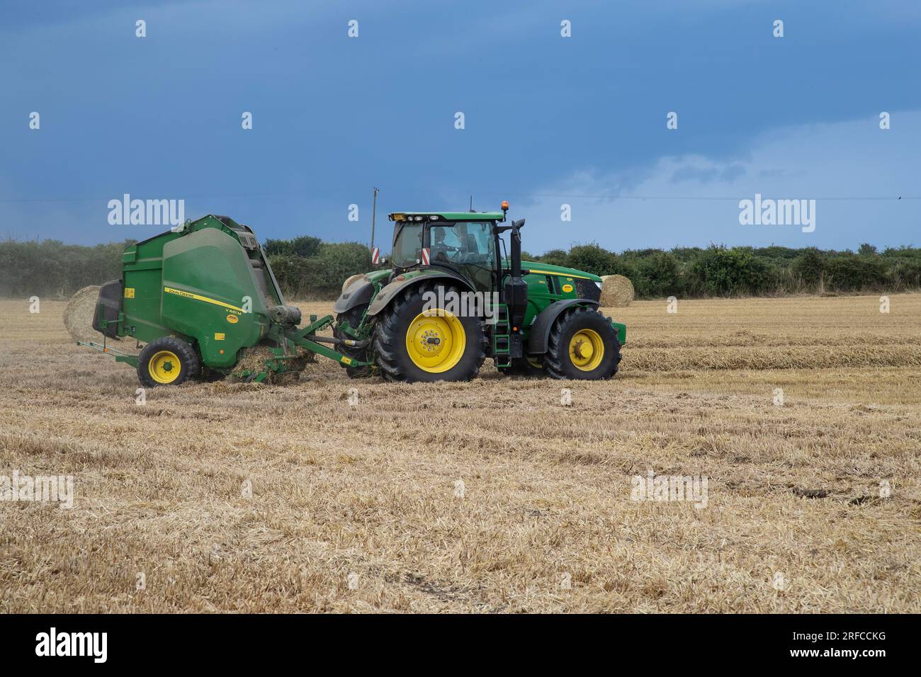 John Deere Tractor and V461M variable chamber baler producing round hay bales in a wheat field during harvesting Stock Photo