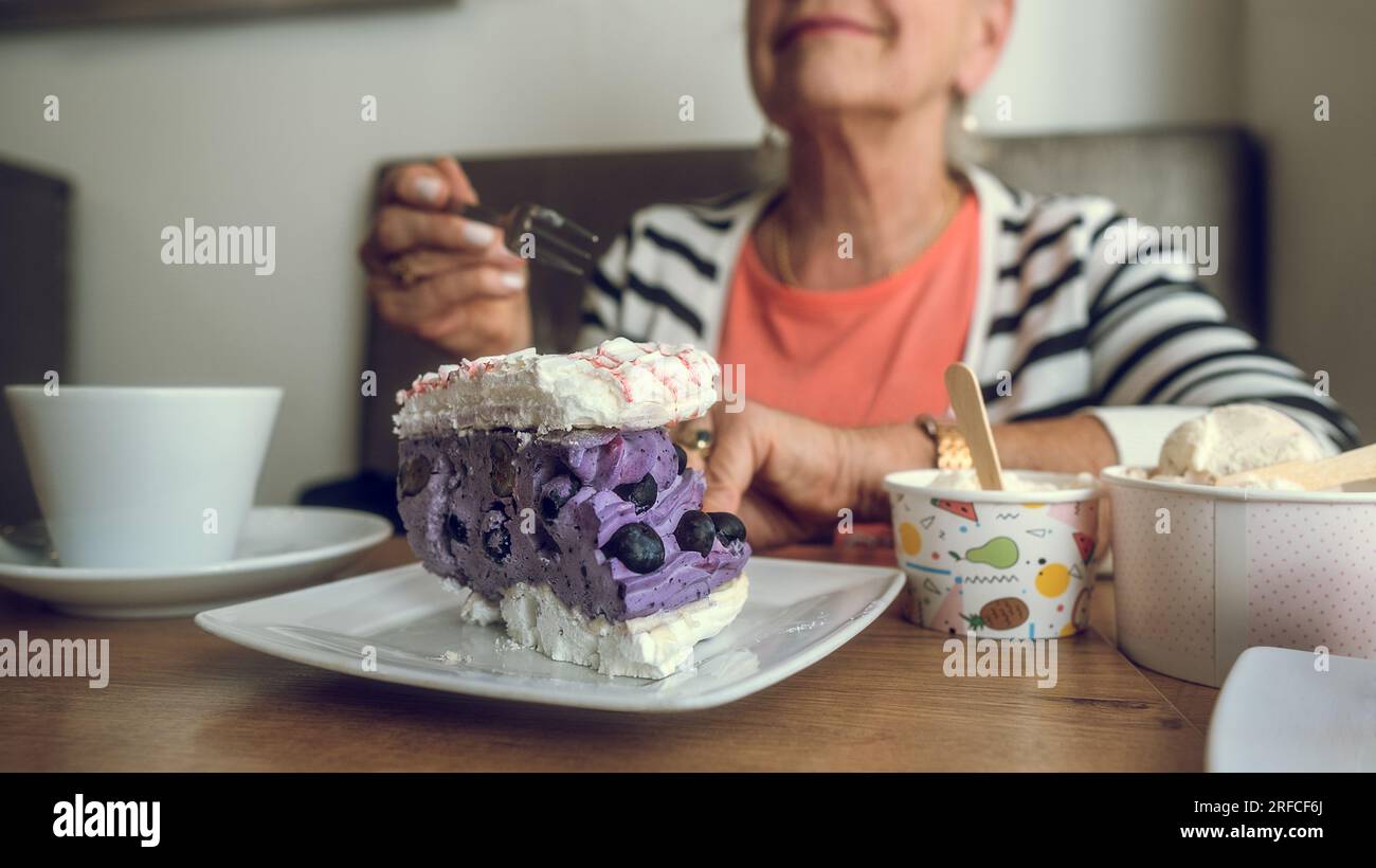 In a cafe. Colorful cake, ice cream and coffee on the table. Hand of an elderly woman with a cake fork. Copy space. Stock Photo