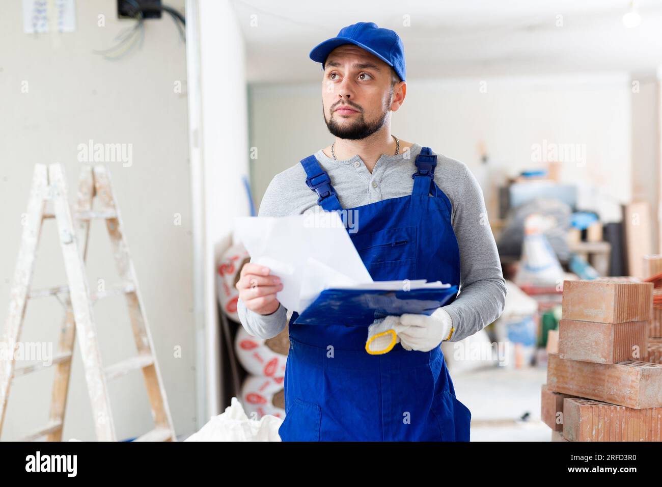 Responsible focused bearded supervisor wearing blue overall taking notes on papers while inspecting construction site Stock Photo