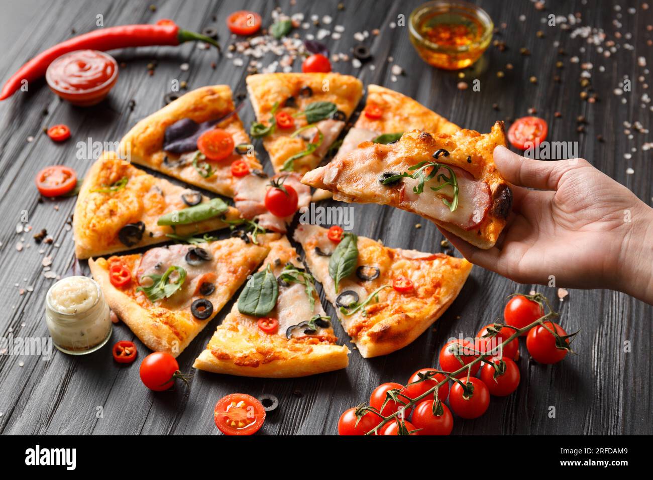 Hands take slices of cut pizza on a dark wooden background top view. Pizza with salami, jerky, sauces, spices, fresh cherry tomatoes on a black Stock Photo