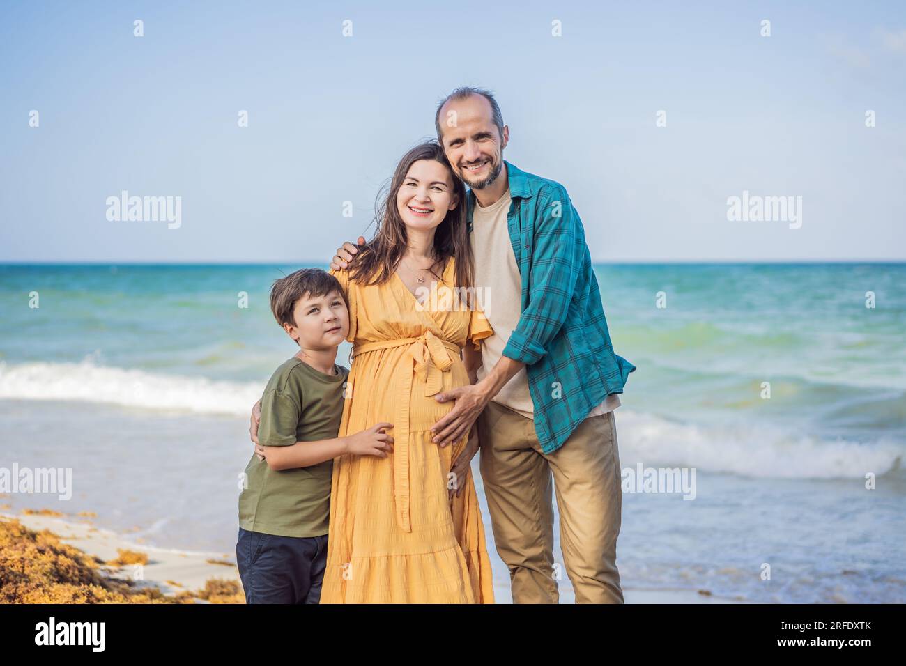 A loving family enjoying tropical beach - a radiant pregnant woman after 40, embraced by her husband, and accompanied by their adult teenage son Stock Photo