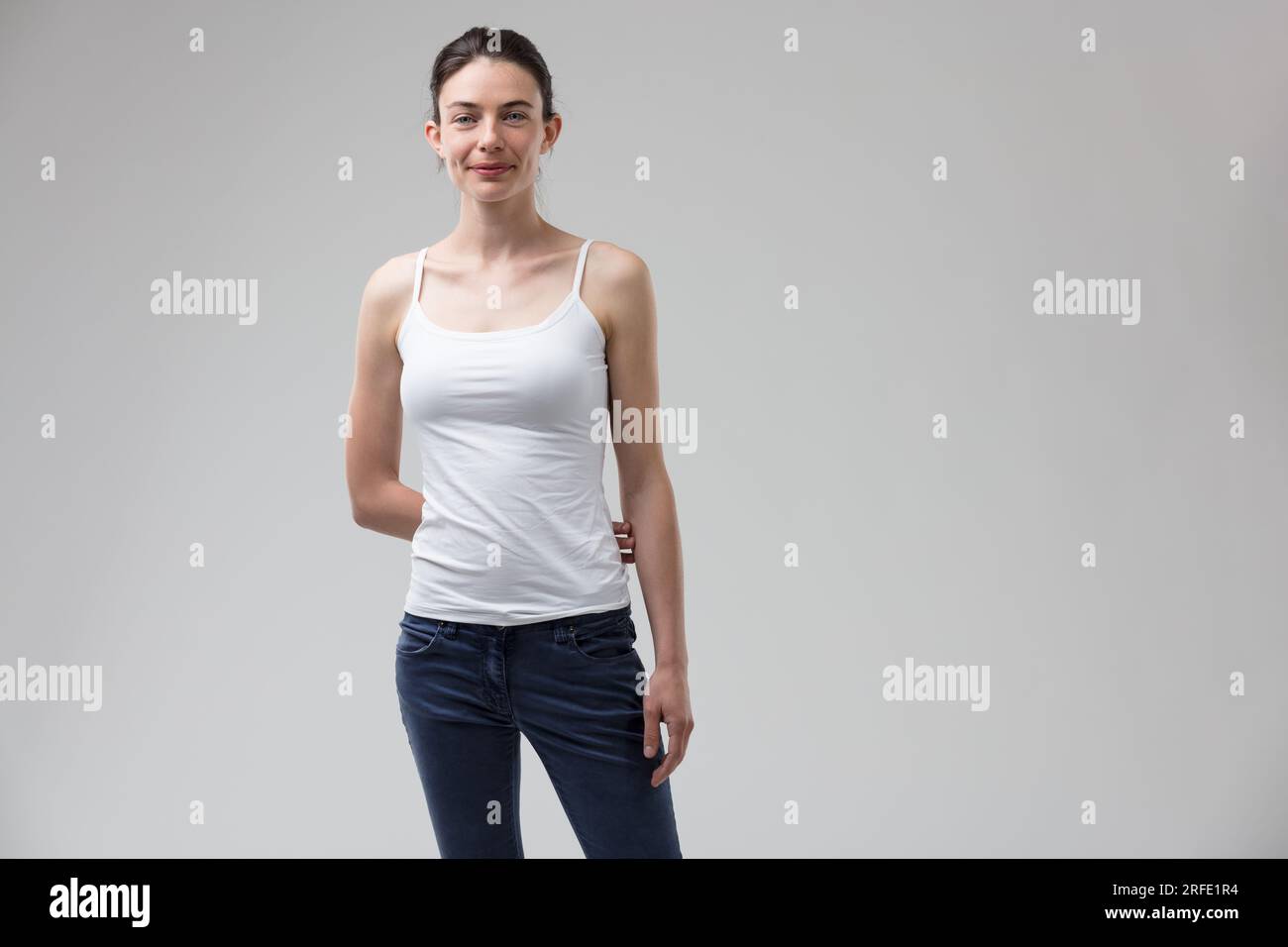 beautiful, slim woman exudes a hint of androgyny or tomboyish charm. With her hair pulled back, wearing a white tank top and blue jeans, she coyly kee Stock Photo