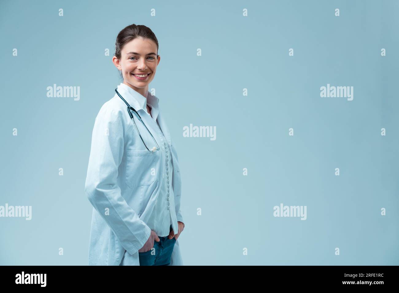 Smiling reassuringly, the satisfied doctor with her lab coat open puts hands in pockets. With light skin and eyes, and tied-up hair, she studied to he Stock Photo