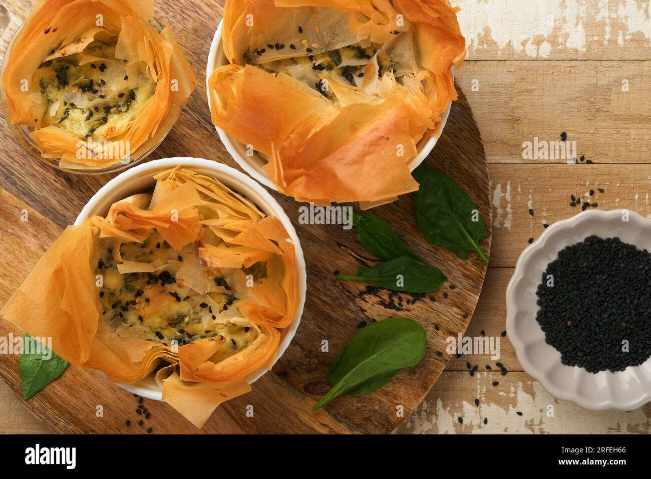 Filo pies with soft feta cheese and spinach in ceramic molds on old wooden table background. Filo portions pies. Small Baked Spanakopita pies. Top vie Stock Photo