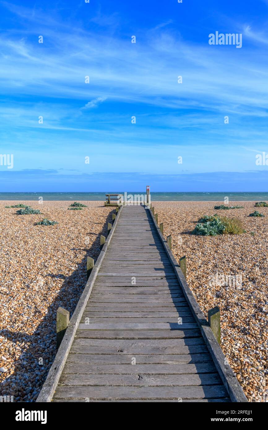Boardwalk with bench on Dungeness beach on SE coast of the UK. Miles of desolate shingle beach, isolated black-painted cottages and a power station! Stock Photo