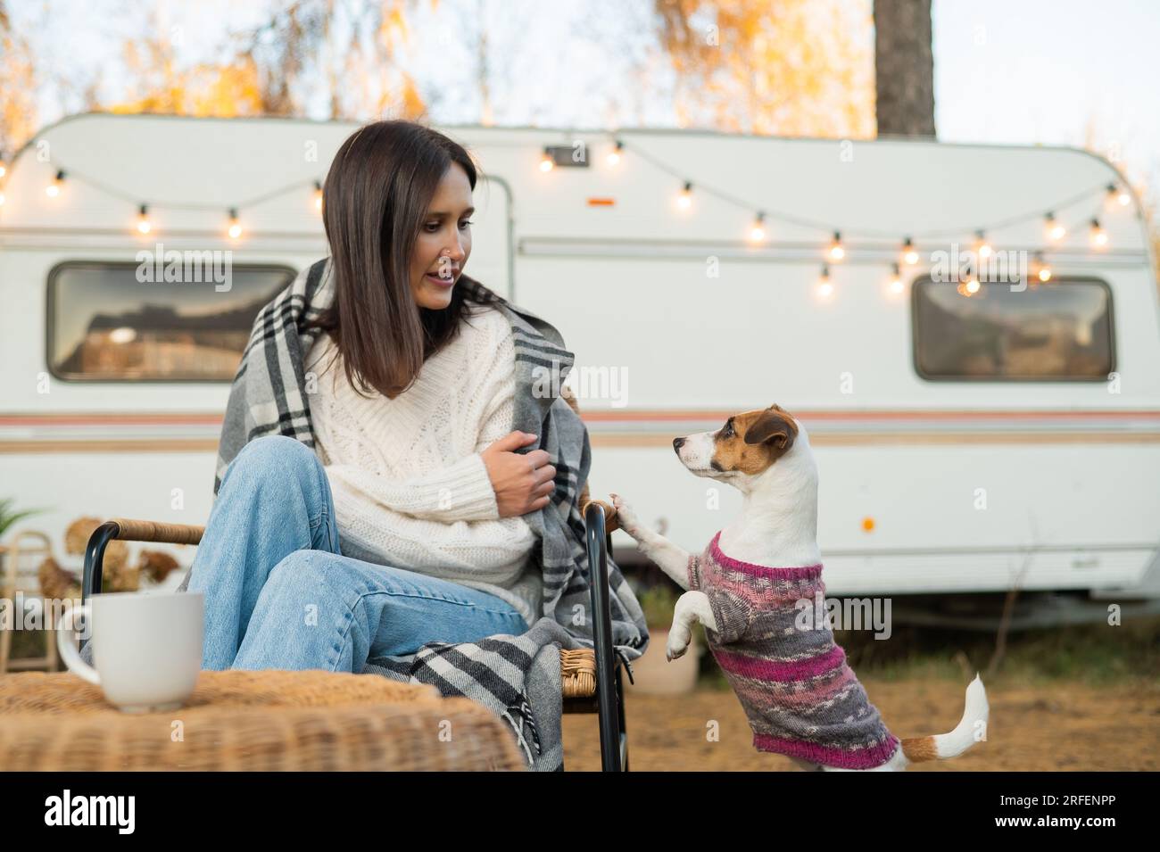 Caucasian woman sitting in a wicker chair wrapped in a blanket with a dog in the yard near the trailer in autumn.  Stock Photo