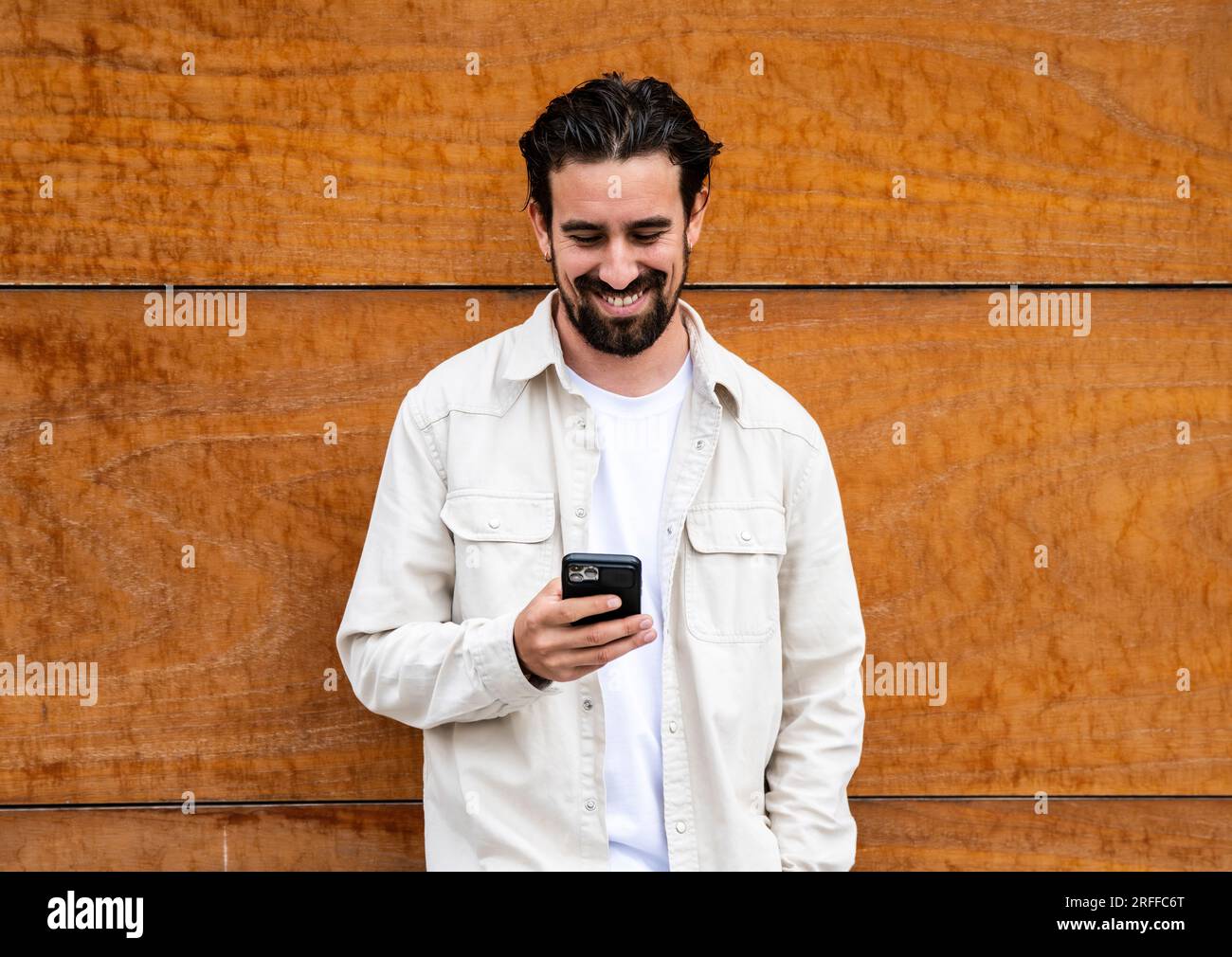 Confident handsome young guy leaning against a wooden wall while texting with his phone. Happy and relaxed man using his smartphone standing on a Stock Photo
