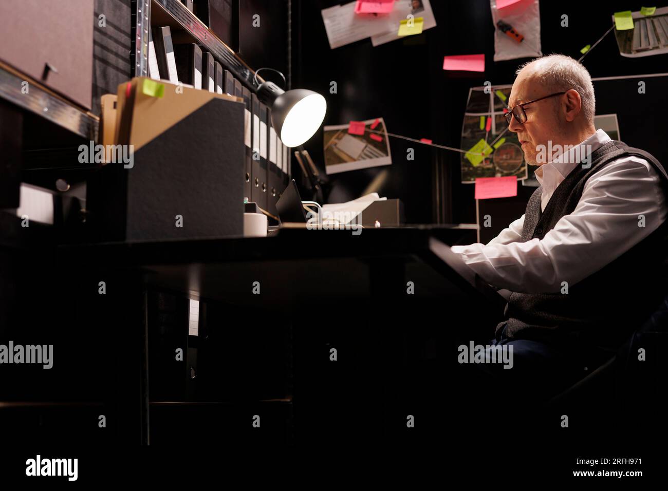 Senior police officer analyzing federal documents, working overtime at criminal case in arhive room. Elderly private detective analyzing mysterious suspect report, checking victim files Stock Photo