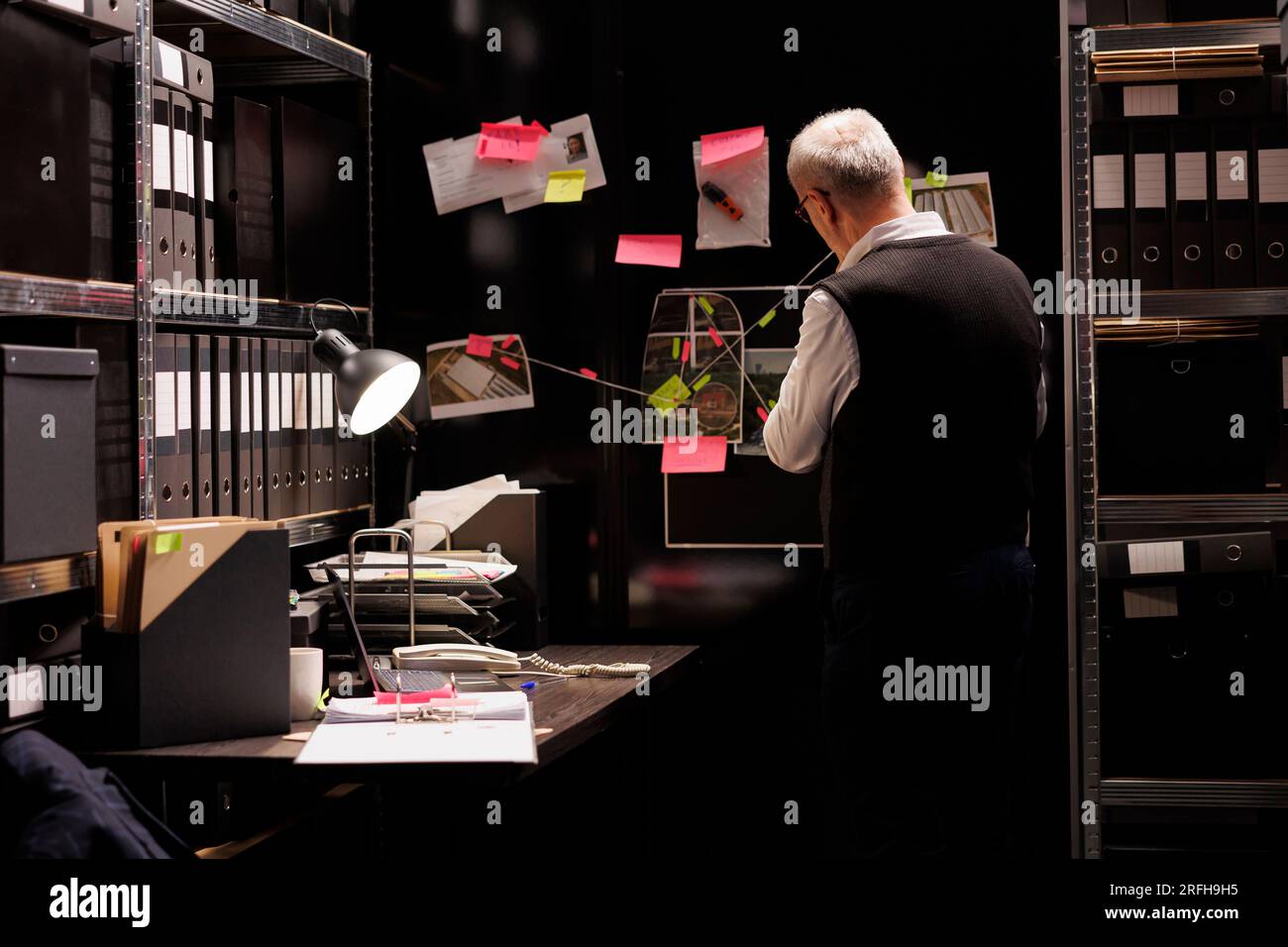 Senior police inspector looking at evidence board, analyzing victim report while working late at night at criminal case in arhive room. Elderly private detective checking criminology files Stock Photo