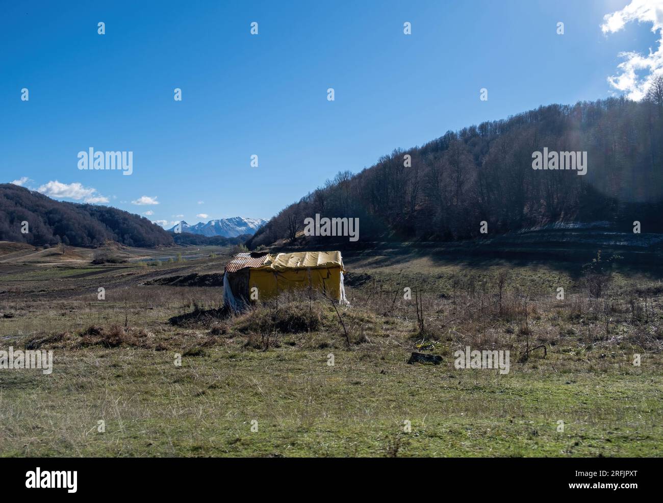 Hut, warehouse in middle of low vegetation field winter sunny day at Aoos Springs Lake region, Epirus Greece. Mountain with forest background. Stock Photo