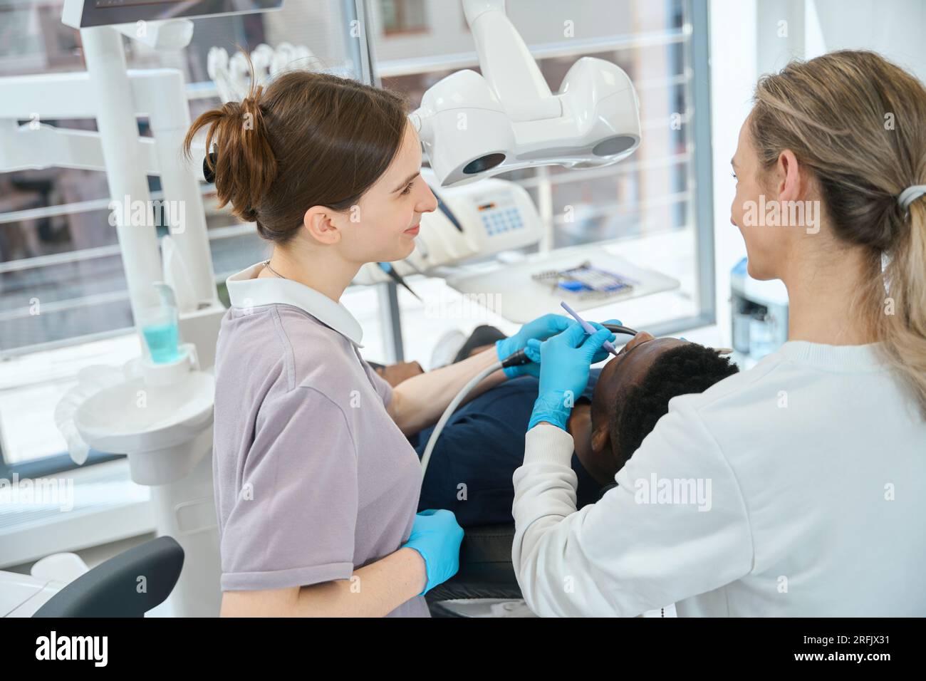 Smiling nurse holding saliva ejector while woman professional dentist keeping root canal therapy or filling teeth to African American client, professi Stock Photo