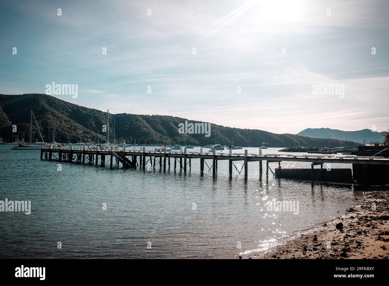 An image of a pier in the ocean. The sky is Blue and clear. In the back ground is boats and hills. Stock Photo