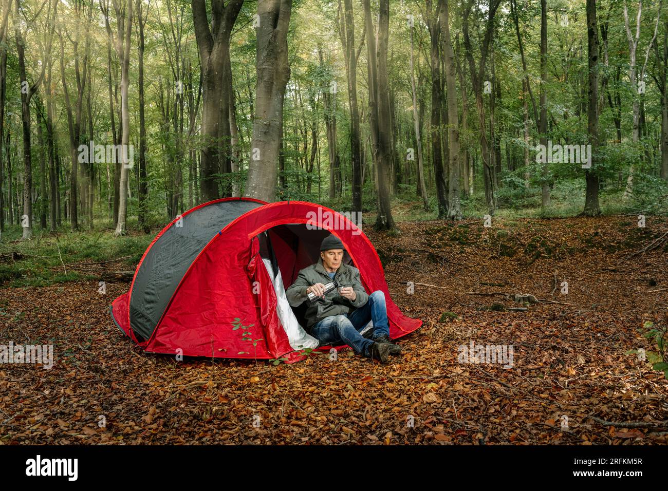 Man sitting in front of his tent in the middle of a forest pouring a coffee from a thermos flask Stock Photo