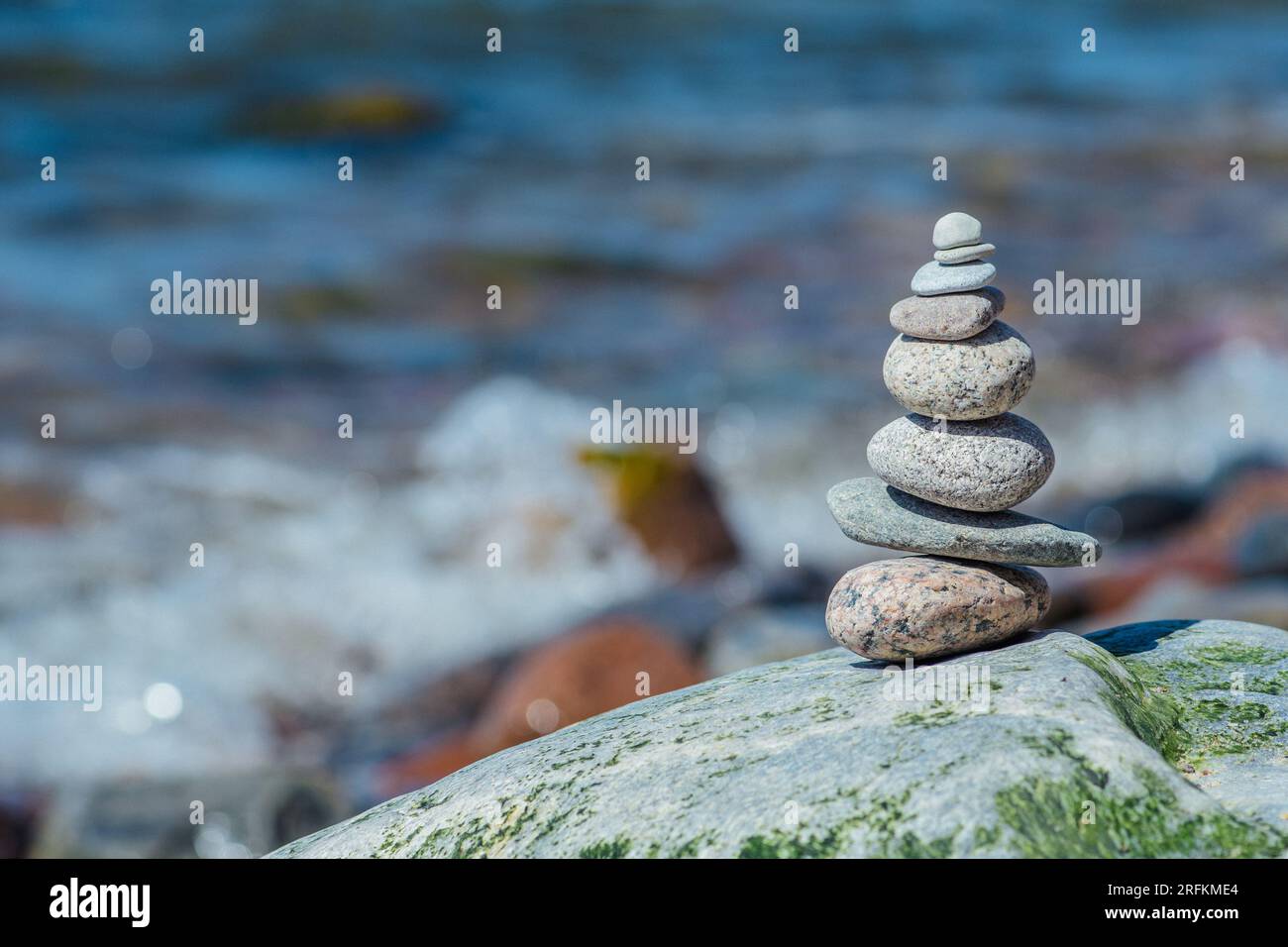 Balancing stones on a big rock with sea grass on the beach, with blue water waves on background, Baltic Sea, Olando Kepure, Lithuania Stock Photo