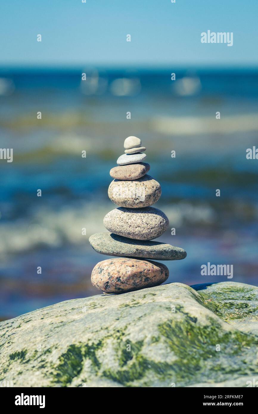 Balancing stones on a big rock with sea grass on the beach, with blue water waves on background, Baltic Sea, Olando Kepure, Lithuania, vertical Stock Photo