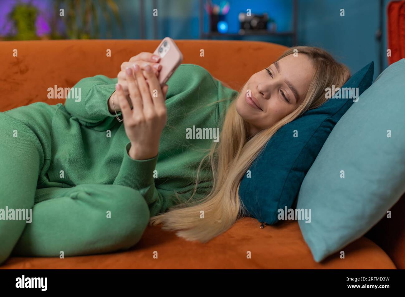 Young woman lying on couch uses mobile phone smiles at home living room