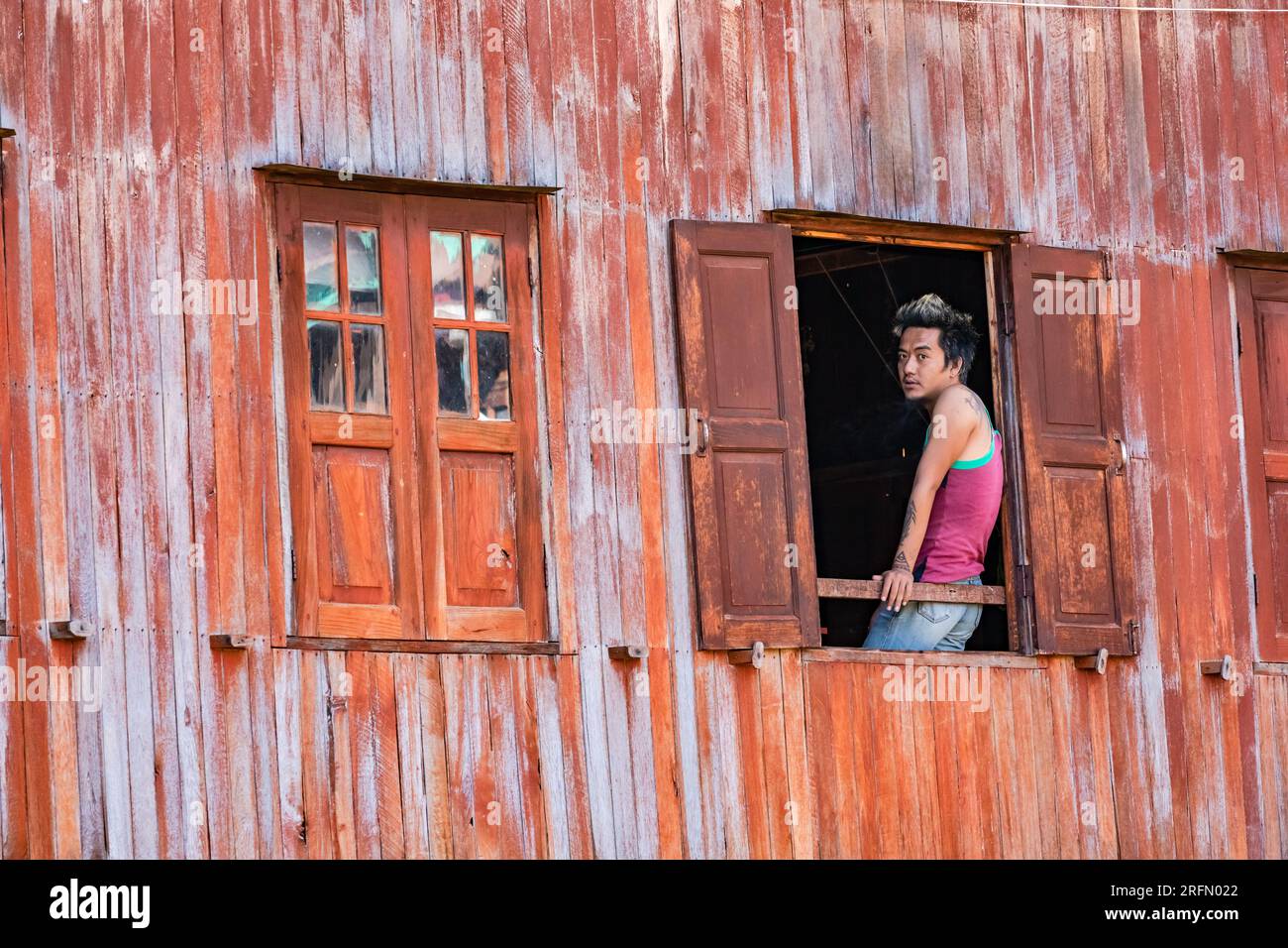 A young man with tatoos and dyed hair looks out of an open window of an orange wooden facade at Inle Lake in Myanmar Stock Photo