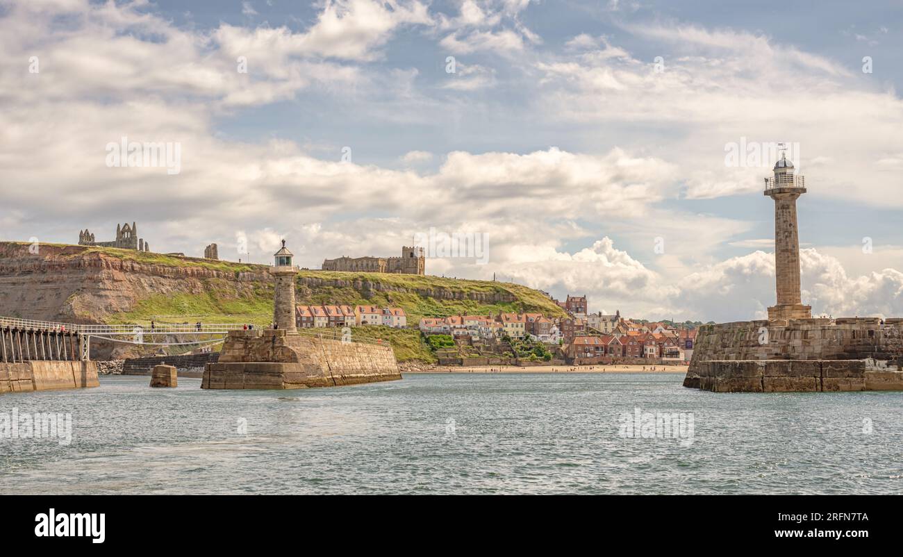 A panorama of Whitby from the sea. A pair of lighthouses guard the entrance to a harbour. The Abbey and a church are on the cliff and people are on a Stock Photo