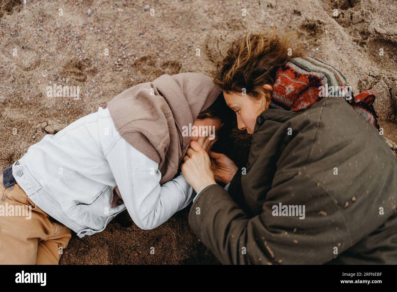 Mother and son quarrel. Kid covered his face with hands and cries lying on the sand. Boy need parent care, mother calms. Stock Photo