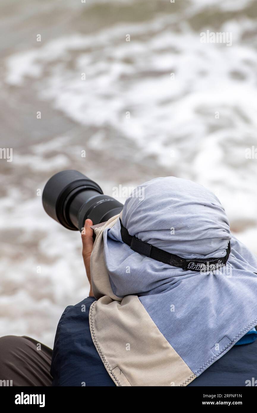 photographer using telephoto long lens to take pictures at the seaside. photographer wearing bandana using long lens. paparazzi photographer. Stock Photo