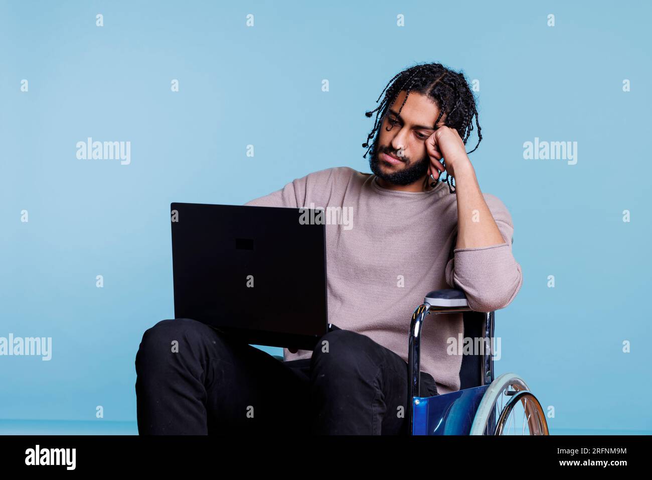 Tired arab man in wheelchair typing on laptop while working remotely. Young exhausted freelancer software developer with physical imparment analyzing code on portable computer Stock Photo