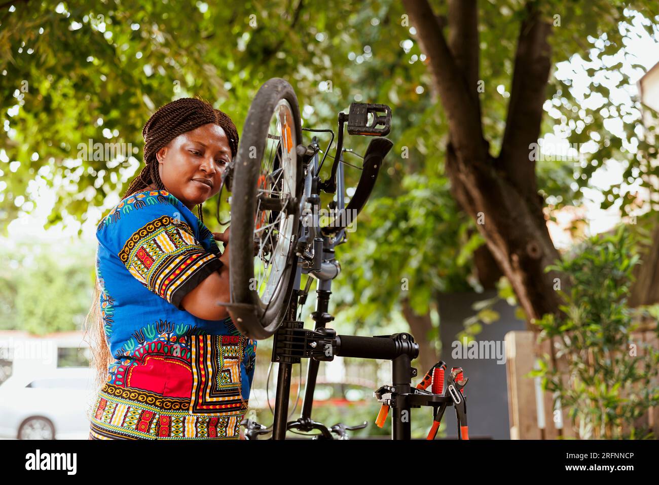 Focused healthy african american woman mending her own bicycle in yard and performing annual outdoor maintenance using expert tools. Female cyclist inspects and repairs bike pedals. Stock Photo