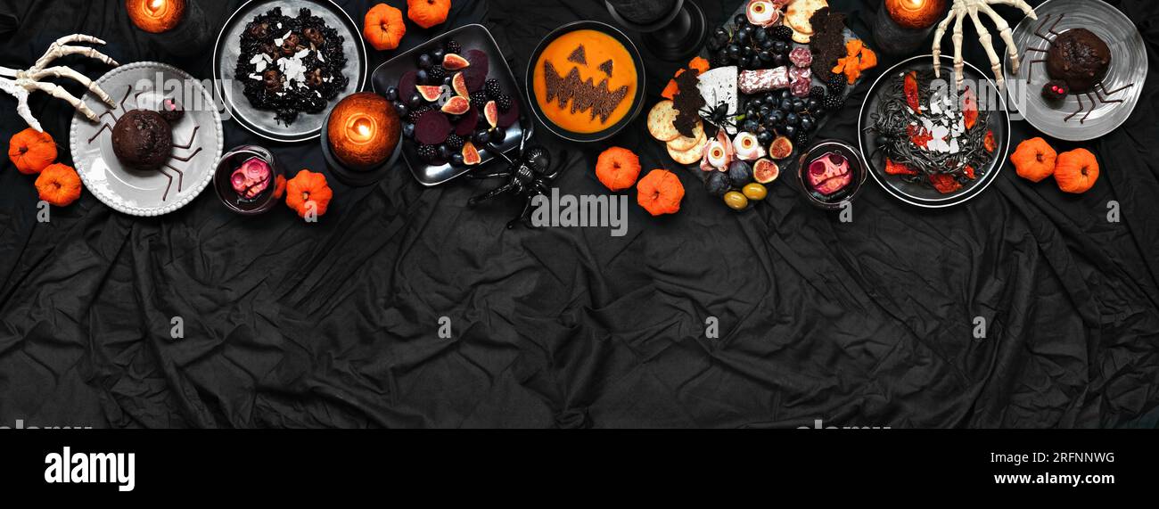 Halloween dinner party top border over a dark black banner background. Above view. Spooky charcuterie board, black risotto and pasta, spider cakes, ja Stock Photo