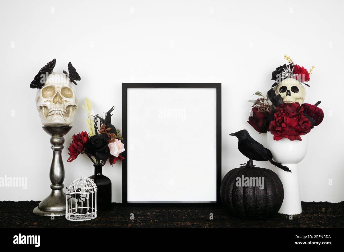 Halloween gothic romance mock up. Black frame on a black shelf with red and black flowers, skulls and pumpkin. Portrait frame against a white wall. Co Stock Photo
