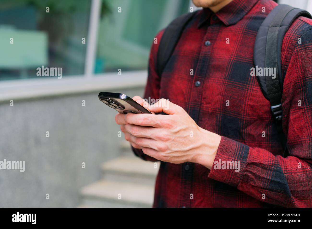 Unrecognizable young modern urban man in red shirt using modern mobile smartphone in hands-on outdoors, 5g internet, using text messages, video calls Stock Photo