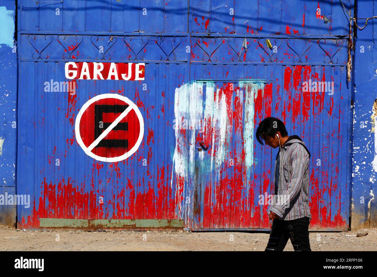 Youth wearing earphones walking past no parking symbol on blue and red painted metal garage doors, El Alto, Bolivia Stock Photo