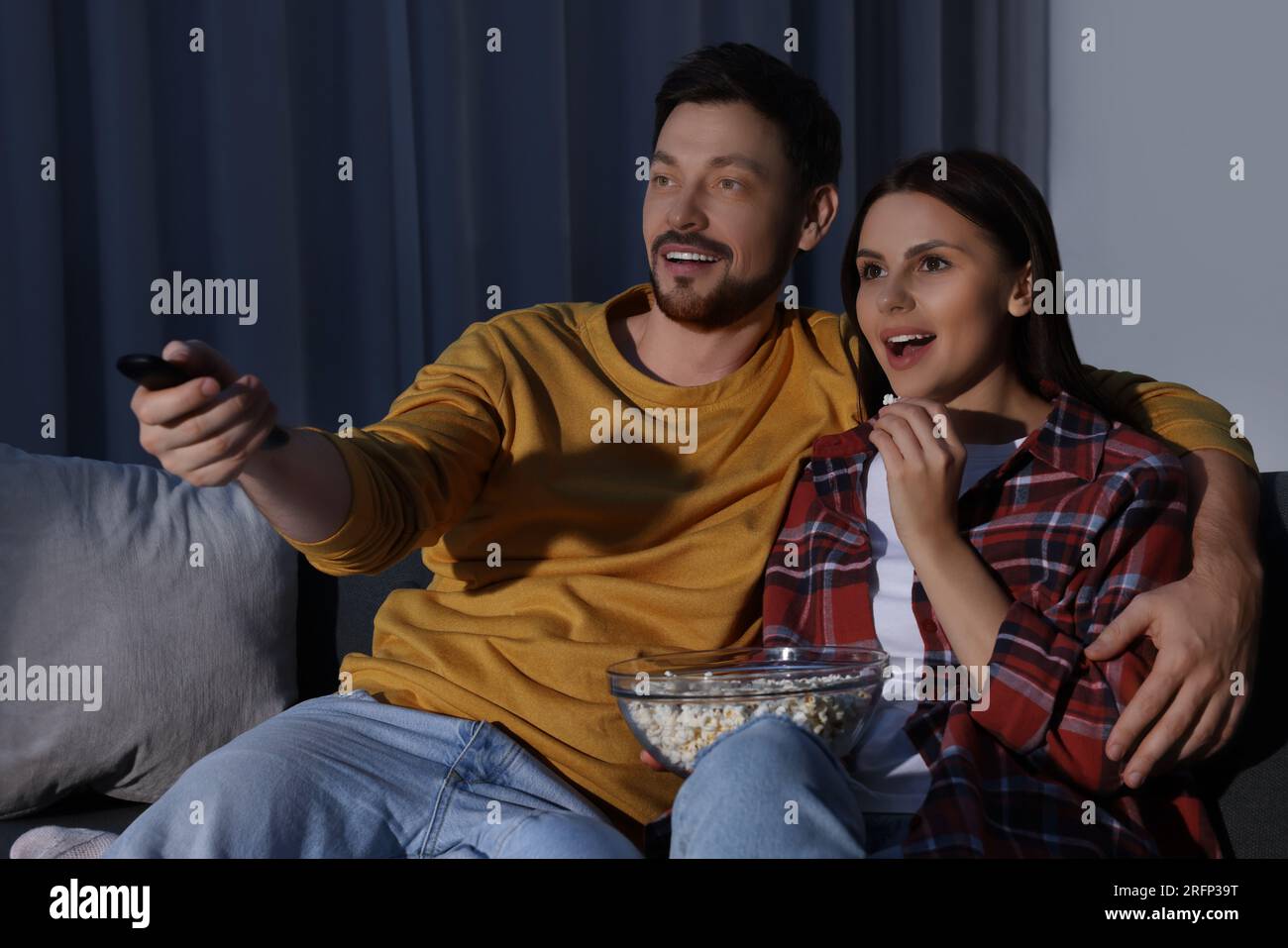 Happy couple watching TV at home in evening. Man changing channels with remote control Stock Photo