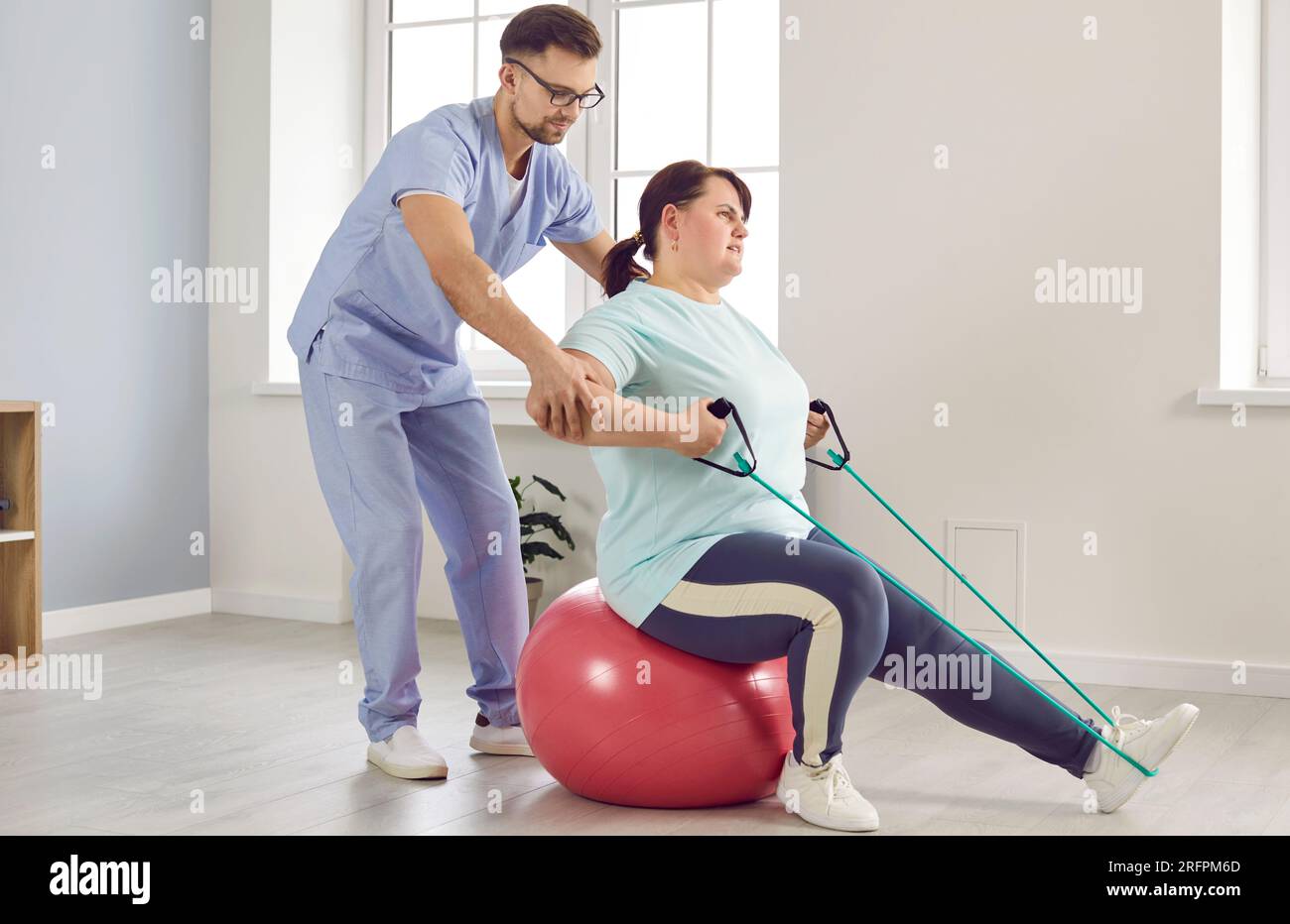 Physiotherapist helps fat woman patient do exercise with fit ball and elastic band Stock Photo