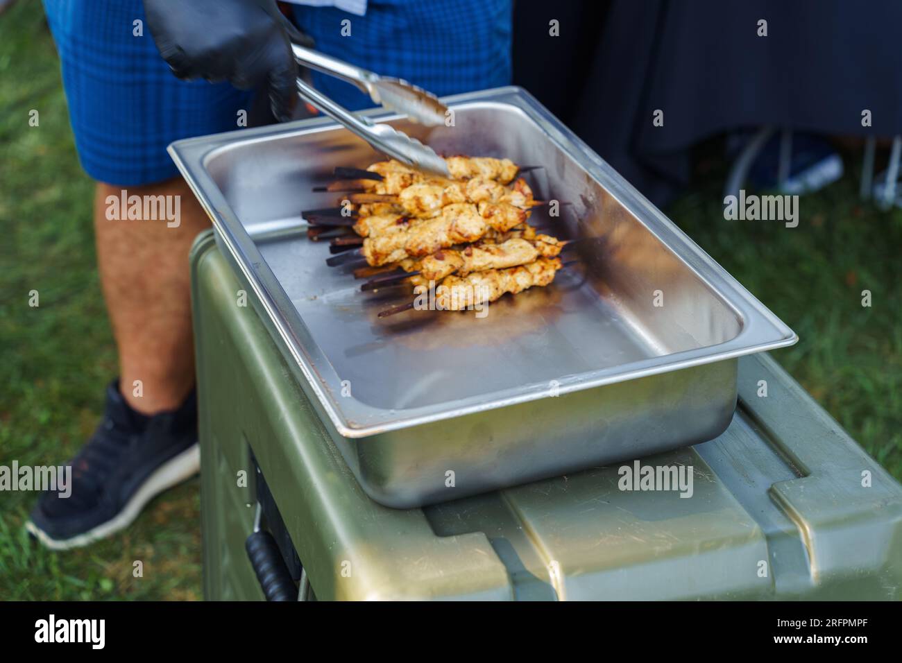 A cook chips grilled chicken kebabs on skewers into a stainless steel container during a barbecue party in the summer Stock Photo