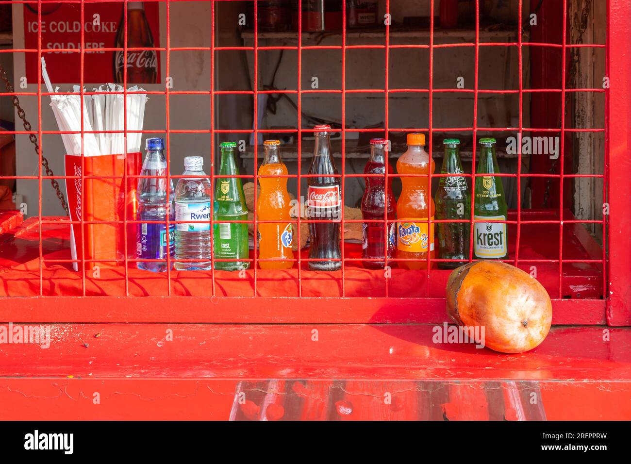 Alignment of bottles offering various drinks behind the fence of a closed red shop. Jinja, Uganda. Stock Photo