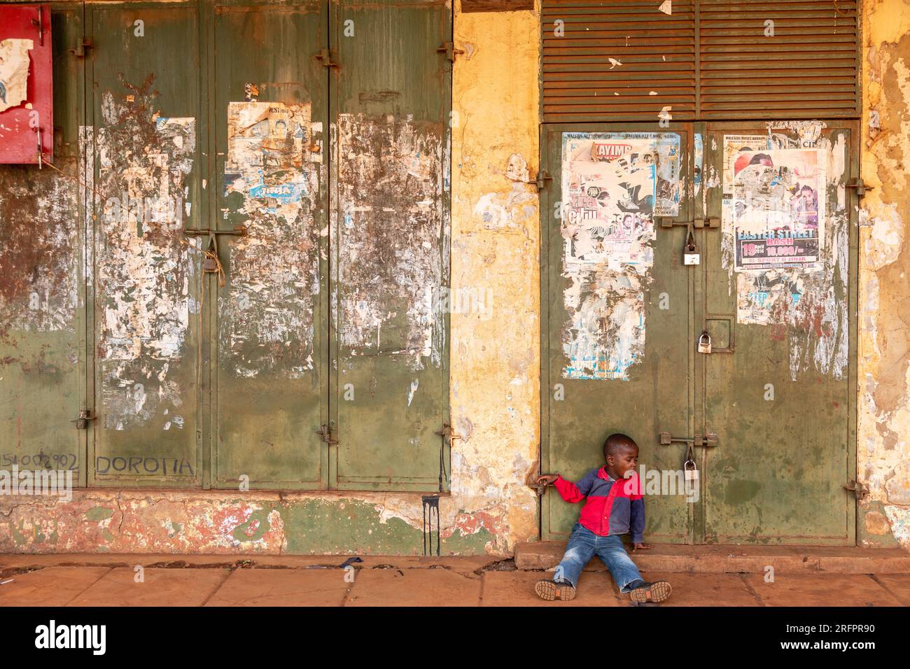 Young boy sitting on the threshold of an iron door. Iron shutters covered with torn posters. Jinja, Uganda. Stock Photo