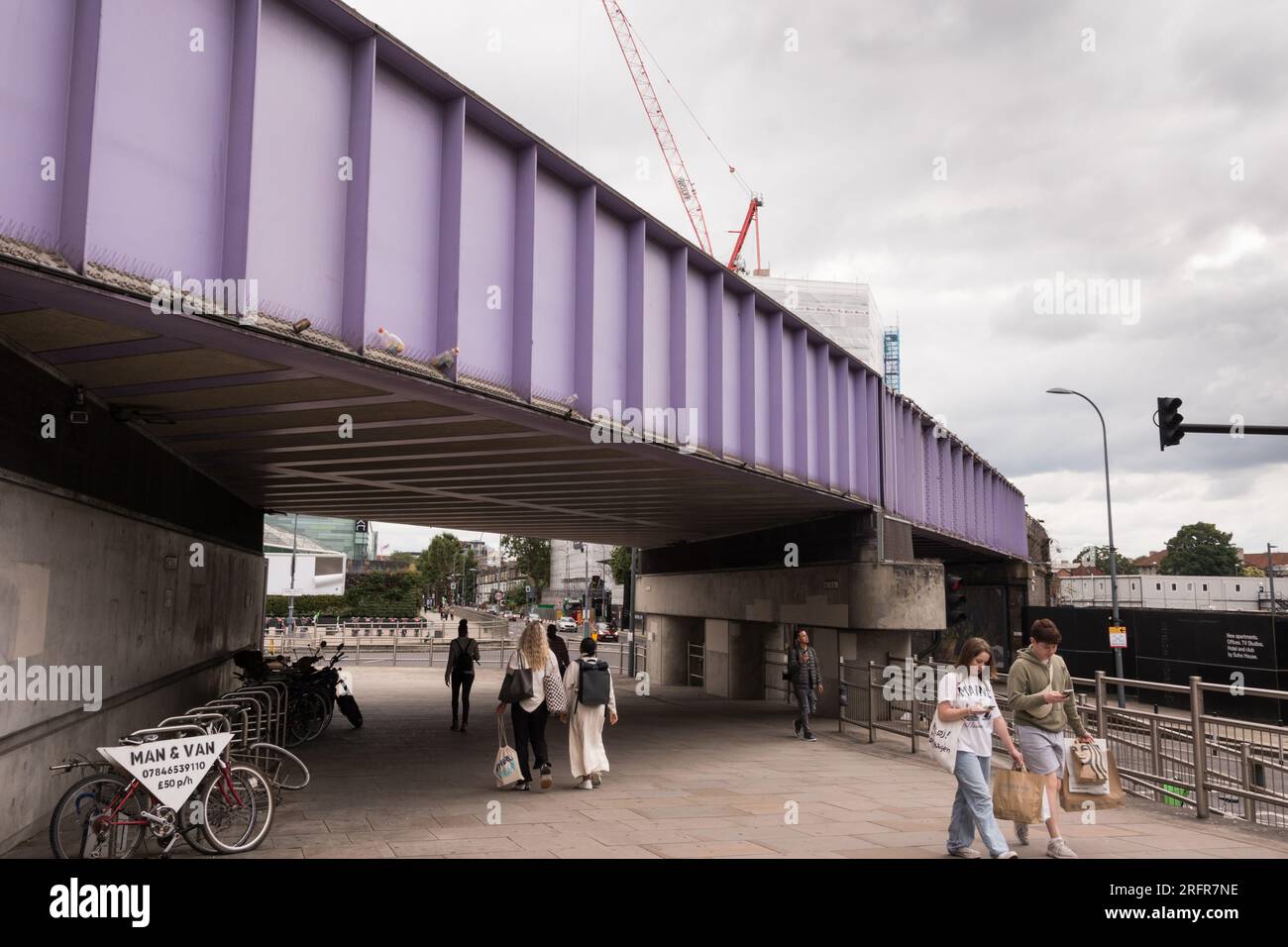 Two young people absorbed in looking at the mobile phones walking under a urple railway bridge next to Wesfield Shopping Centre, London, England, U.K. Stock Photo