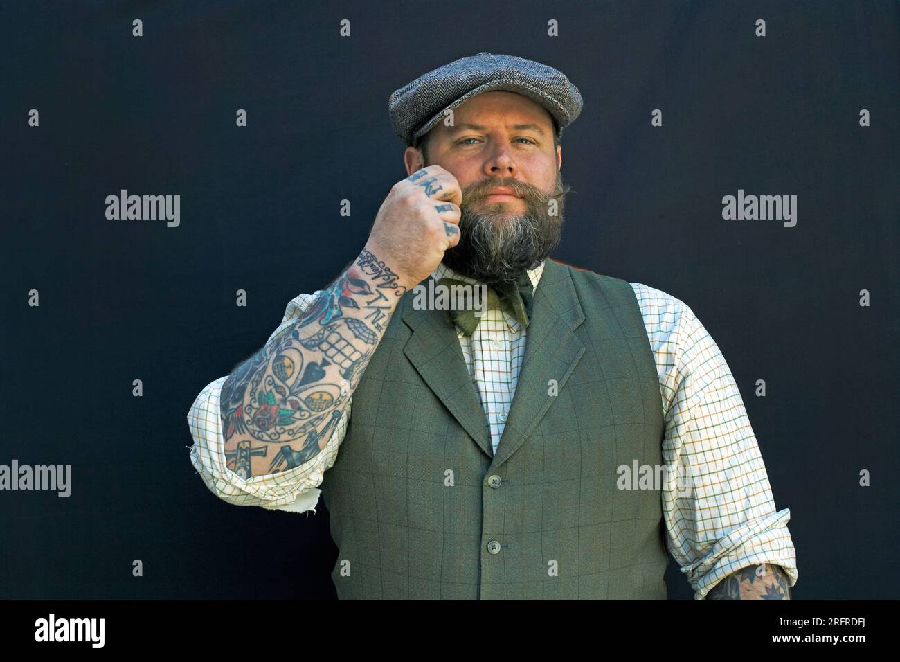 Bearded man is twirling his mustache with his fingers. Portrait of a hipster on black background closeup male with tattoos on his arm. Stock Photo