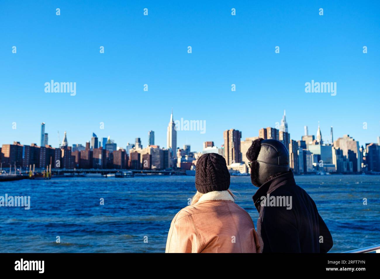 Sunlit couple in winter attire, gazing at vibrant Midtown Manhattan from East River's edge Stock Photo