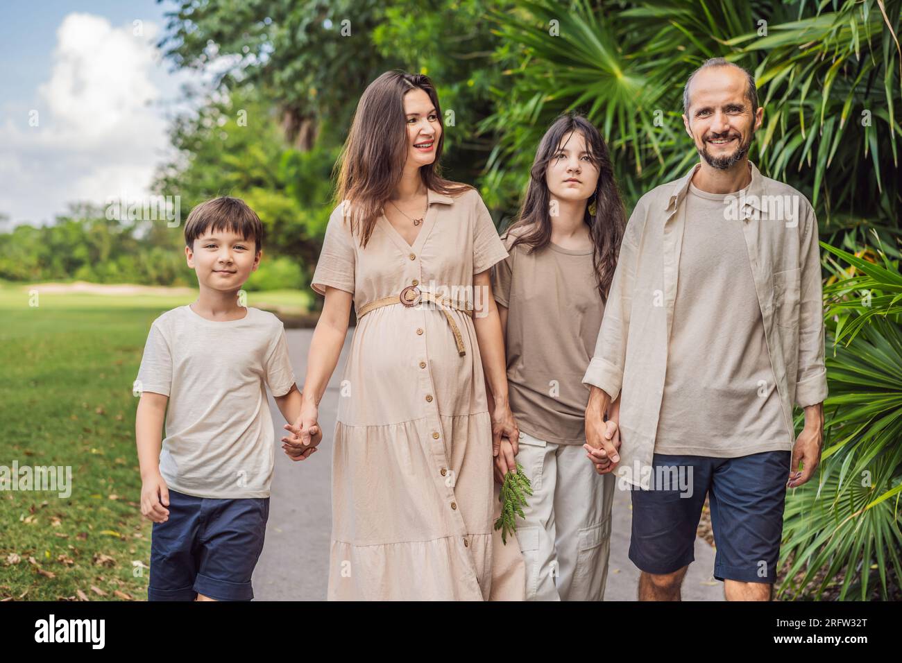 A loving family enjoying a leisurely walk in the park - a radiant pregnant woman after 40, embraced by her husband, and accompanied by their adult Stock Photo
