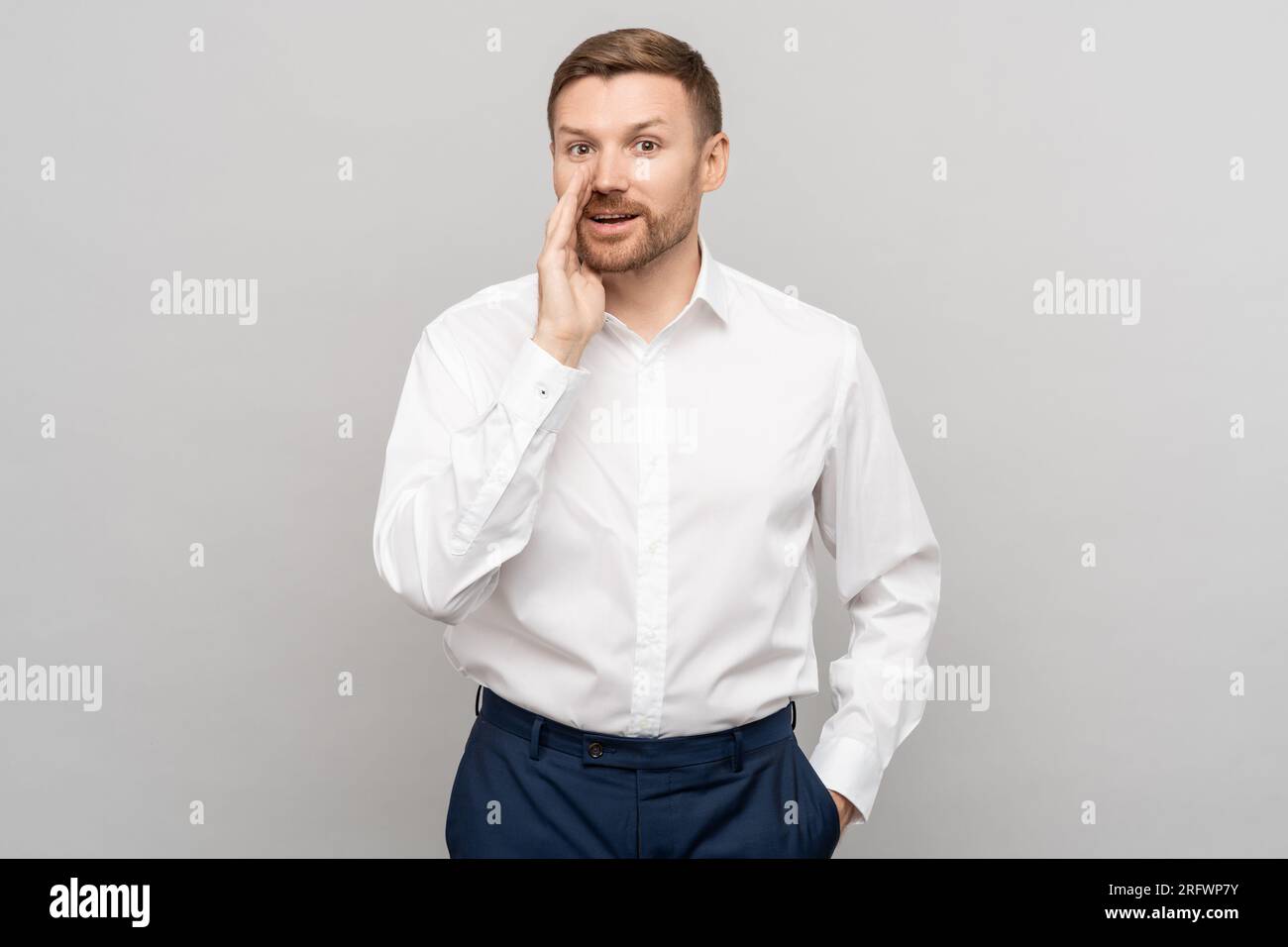 Office worker man shares a secret offer, whispering with hand near mouth gesture on grey background. Stock Photo