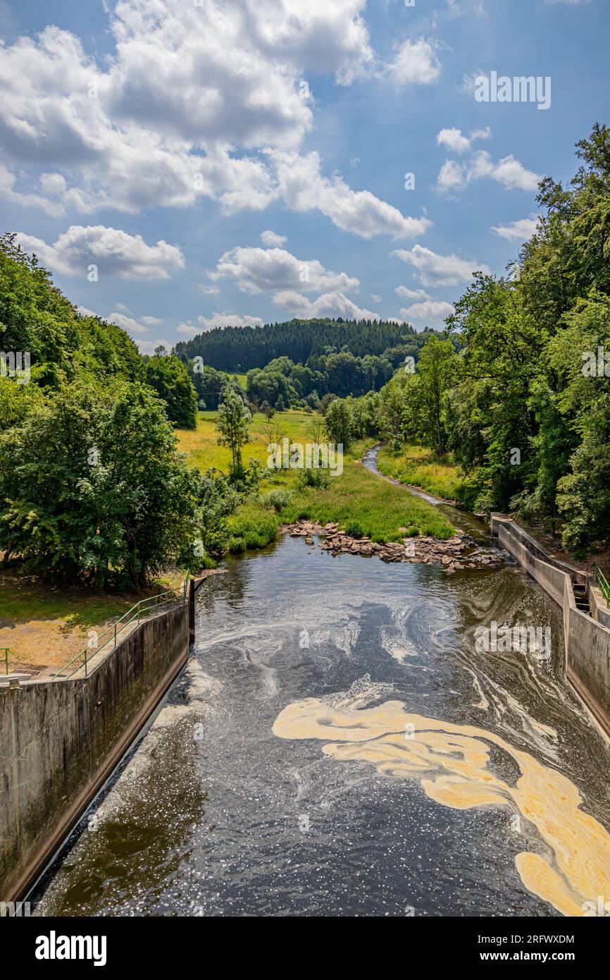 Water flowing out of Staumauer Bitburger dam into Prum river, valley and mountains with leafy trees in background, brown stain on water surface, Staus Stock Photo