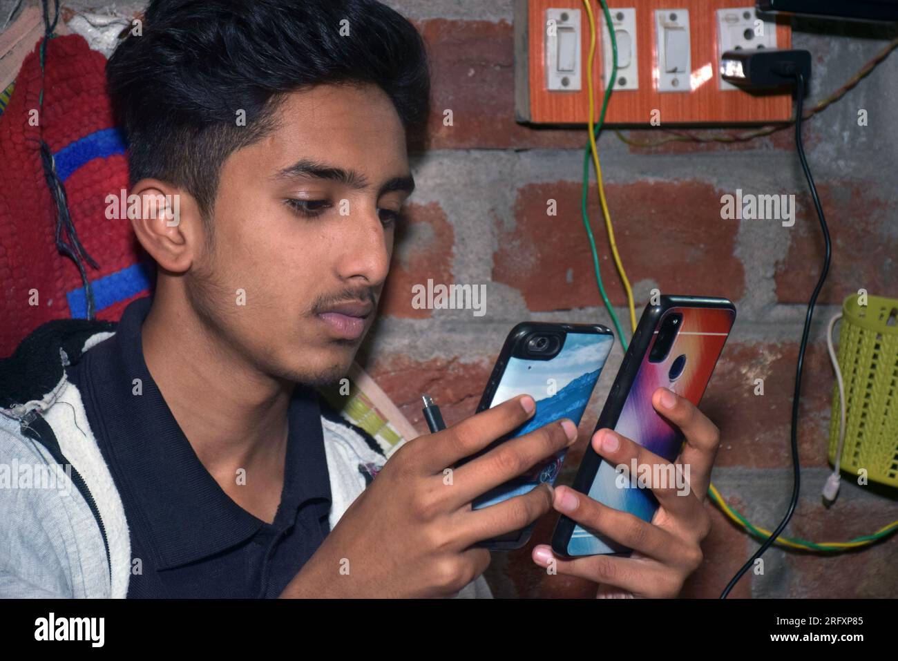 Indian teen using two mobiles simultaneously at home. brick wall and electric board background. Stock Photo