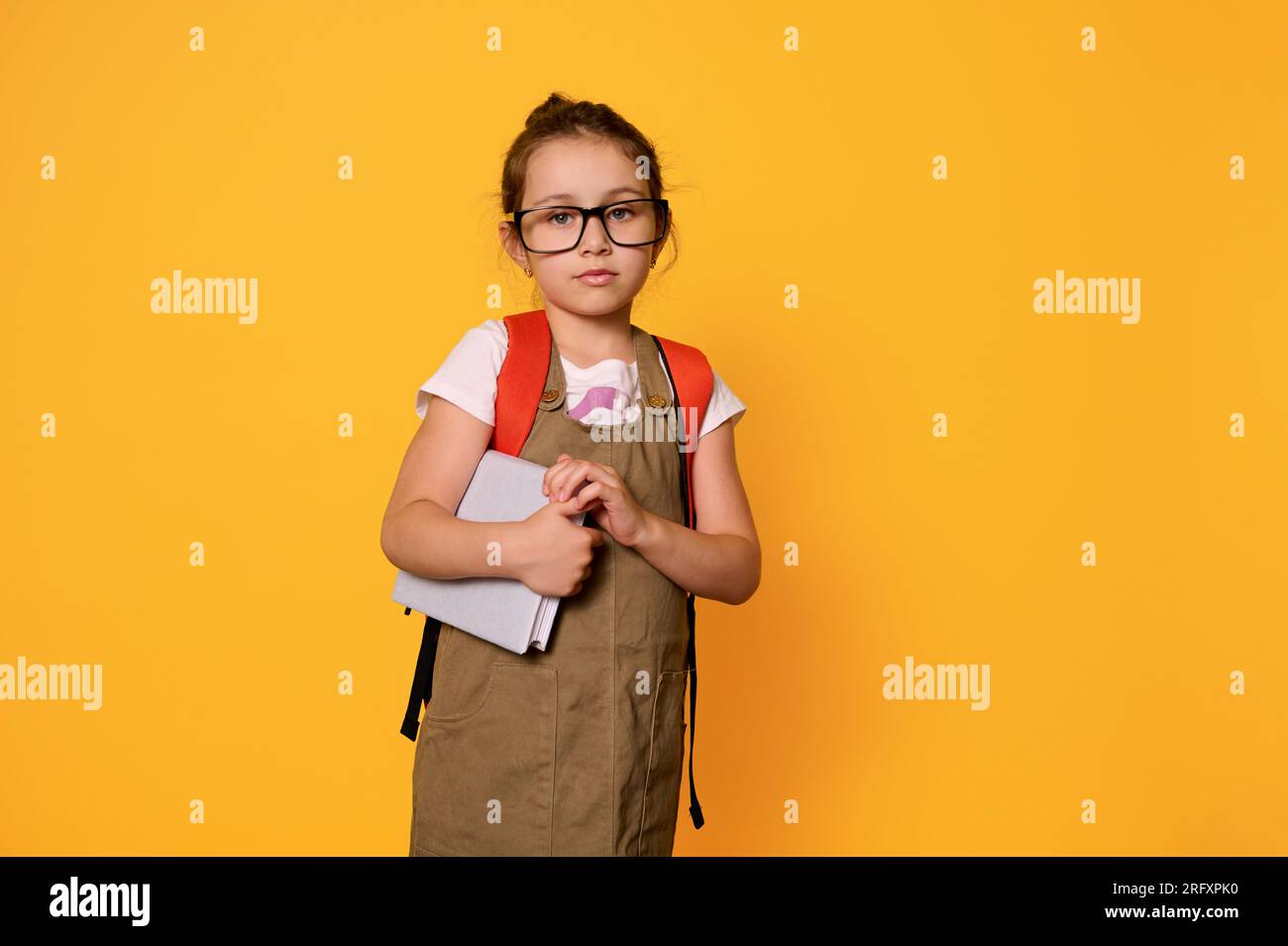 Adevrtising studio portrait of Caucasian confident serious cute little girl 6 years old, with orange backpack, holding a book, dressed in casual cloth Stock Photo