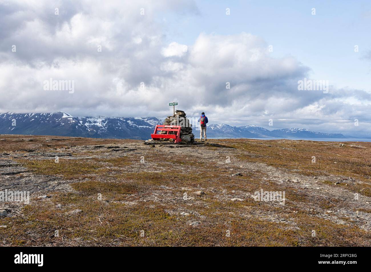 A man with a backpack is standing on the top of Barheia near a big pile of stones and a red bench.Svensby, Lyngen Alps, Lyngsalpan, Northern Norway Stock Photo